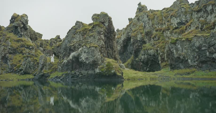 A woman in a white dress stands amidst a dramatic, moss-covered rocky landscape reflected in a still lake