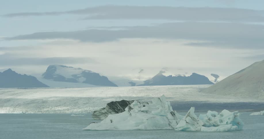 A majestic panning view of icebergs floating serenely in a glacial lagoon, with a vast glacier and mountains in the background
