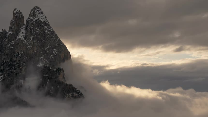 A dramatic time-lapse of a snow-capped mountain peak emerging from a sea of rolling clouds under a moody, overcast sky