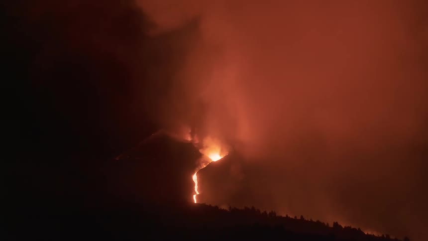 A powerful volcanic eruption at night sends molten lava flowing down the mountainside while thick smoke fills the sky