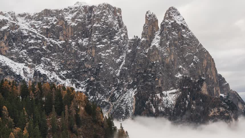 A dramatic timelapse captures clouds swirling around snow-dusted mountain peaks towering over a colorful autumn forest
