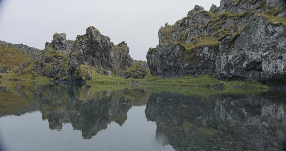 A serene lake reflects mossy rock formations on an overcast day as light rain creates ripples on the surface