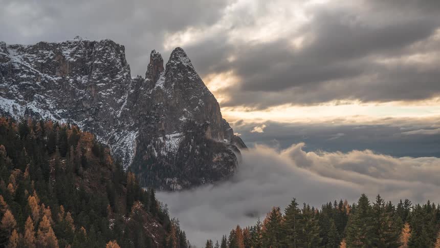 A dramatic timelapse shows a sea of clouds and fog rolling through a mountain valley, surrounding a snow-capped peak at sunrise