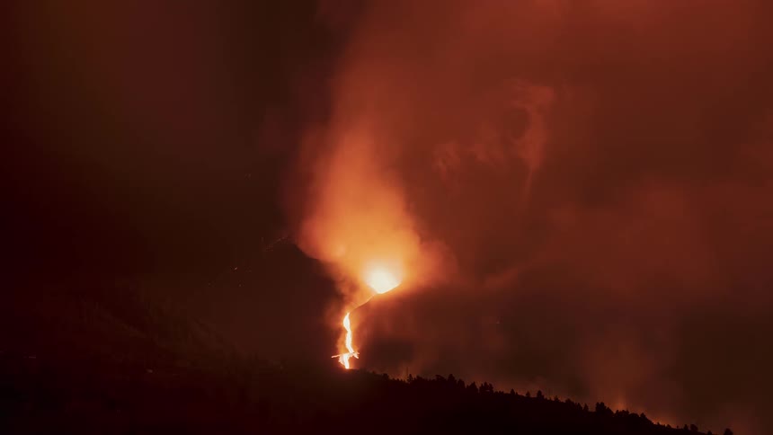 A powerful volcanic eruption at night sends rivers of molten lava down its slopes, illuminating thick plumes of smoke in an ominous red glow