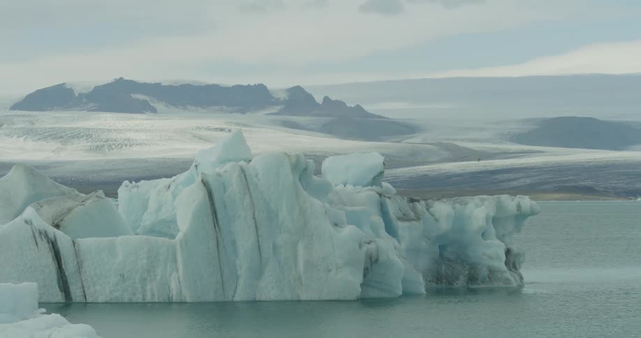 A blurry shot slowly focuses on a massive iceberg floating in a tranquil glacial lagoon with a vast, snowy landscape in the background
