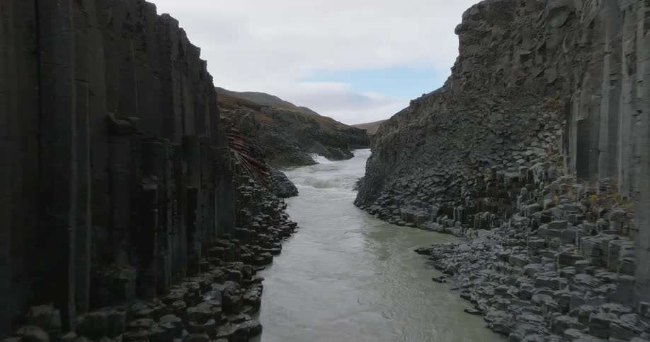 An aerial shot slowly travels through a dramatic canyon lined with unique basalt column rock formations as a river flows below