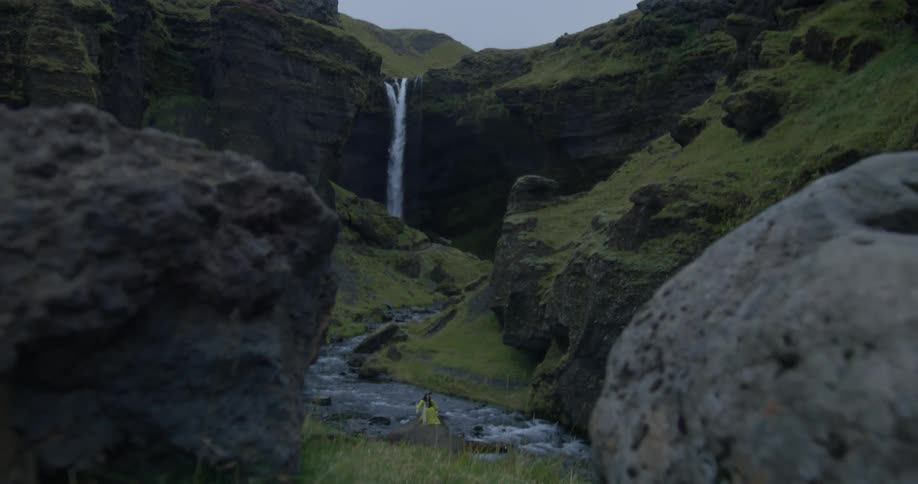 A lone traveler in a bright yellow jacket sits beside a river, admiring a majestic waterfall cascading through a lush green canyon
