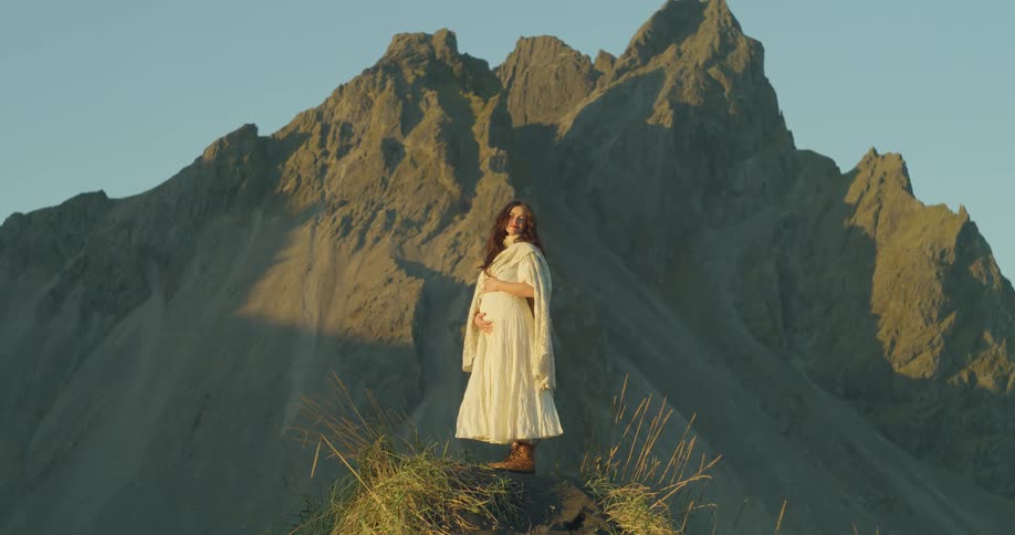 A pregnant woman in a flowing white dress gently holds her belly while standing before a majestic, sunlit mountain