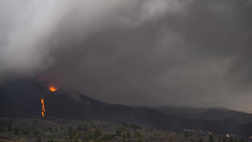Thick dark clouds of smoke and ash billow from an erupting volcano as lava flows down its side