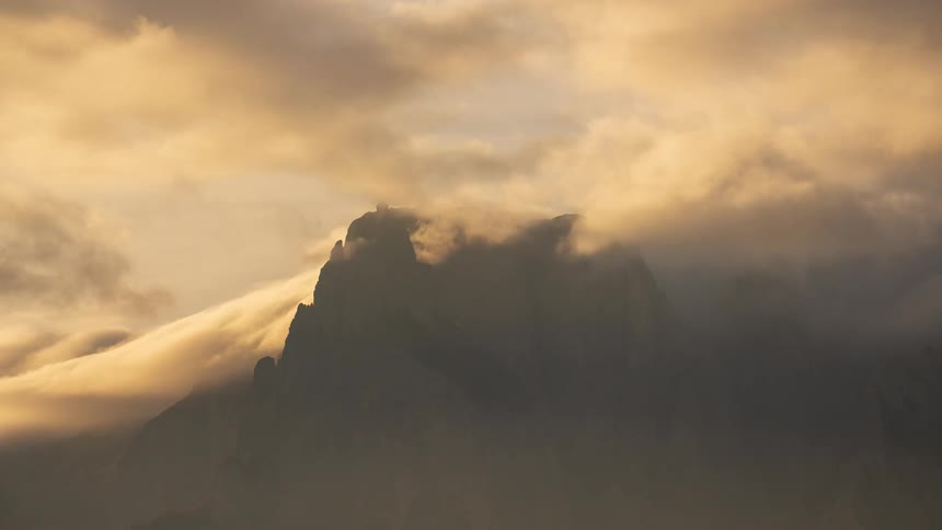 A dramatic timelapse shows golden clouds and thick fog swirling around a rugged mountain peak at sunrise
