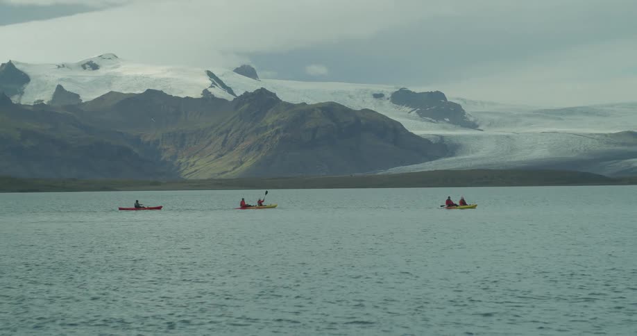 A group of kayakers paddle across a vast lake at the base of a majestic, snow-covered mountain range and glacier
