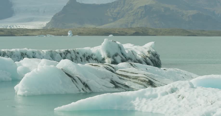 Kayakers paddle across a vast glacial lagoon filled with massive icebergs and a mountain range in the background