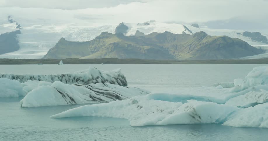 A slow pan across a serene glacial lagoon reveals massive icebergs floating in front of distant snow-capped mountains in Iceland