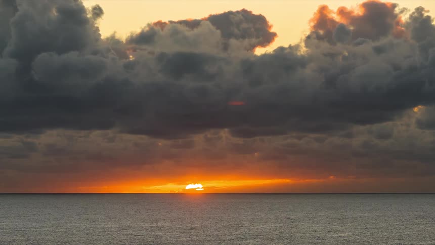 A dramatic timelapse captures the sun rising over the calm ocean, breaking through a thick layer of dark clouds