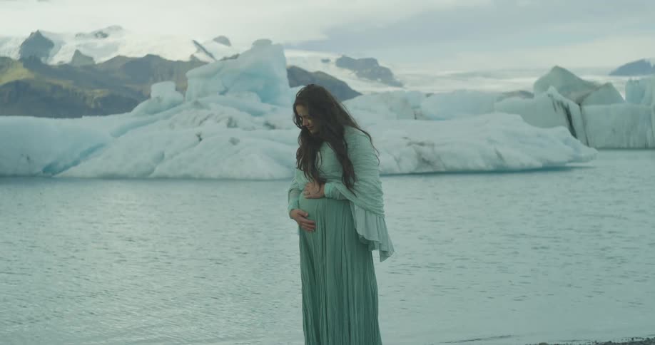 A serene pregnant woman in a flowing green dress lovingly holds her belly on the shore of a glacial lagoon in Iceland