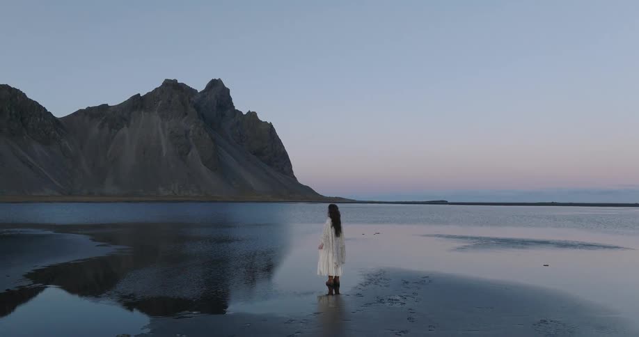 Aerial view pulls back from a woman in a white dress standing on a serene black sand beach with a majestic mountain reflected in the water at dusk