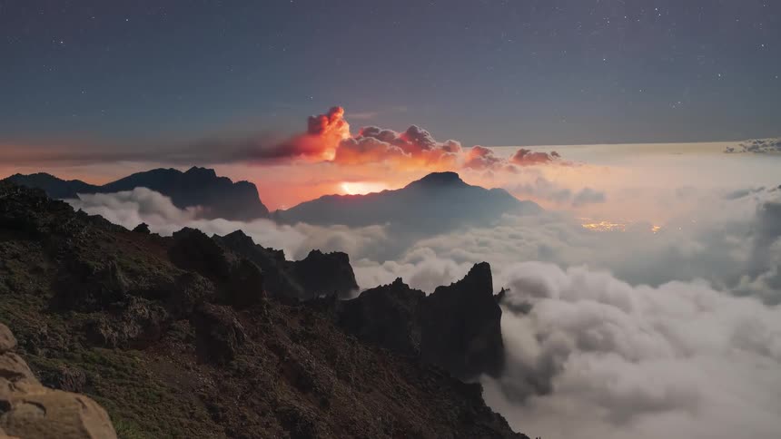A breathtaking day-to-night timelapse from a mountain peak shows clouds flowing through a valley as a distant volcanic eruption illuminates the clouds and sky with a fiery red glow