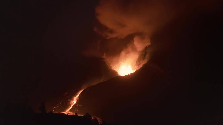 A volcano violently erupts at night, spewing fountains of lava and plumes of smoke into the dark sky as a molten river flows down its slope