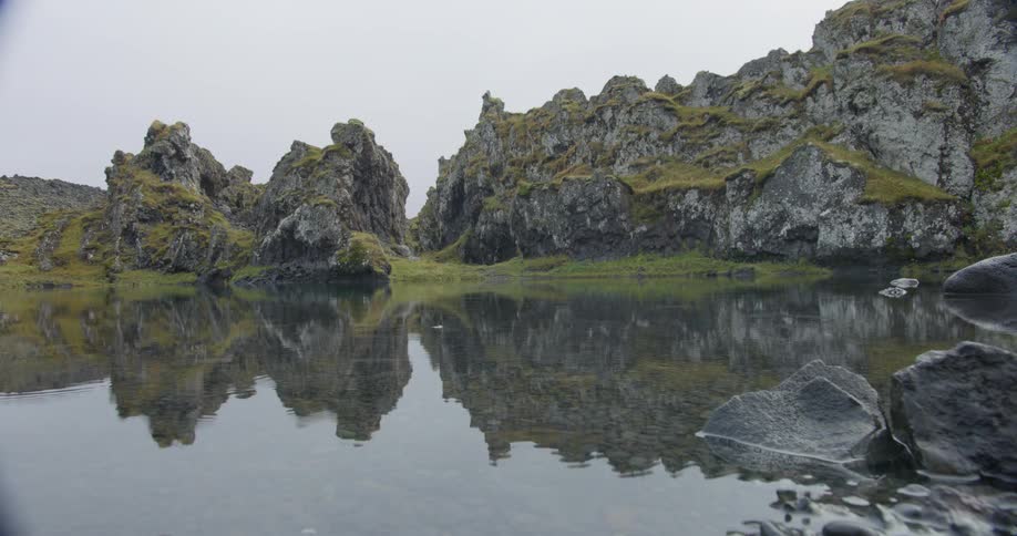 Rugged, mossy rock formations are perfectly reflected in a tranquil lake on a still, overcast day