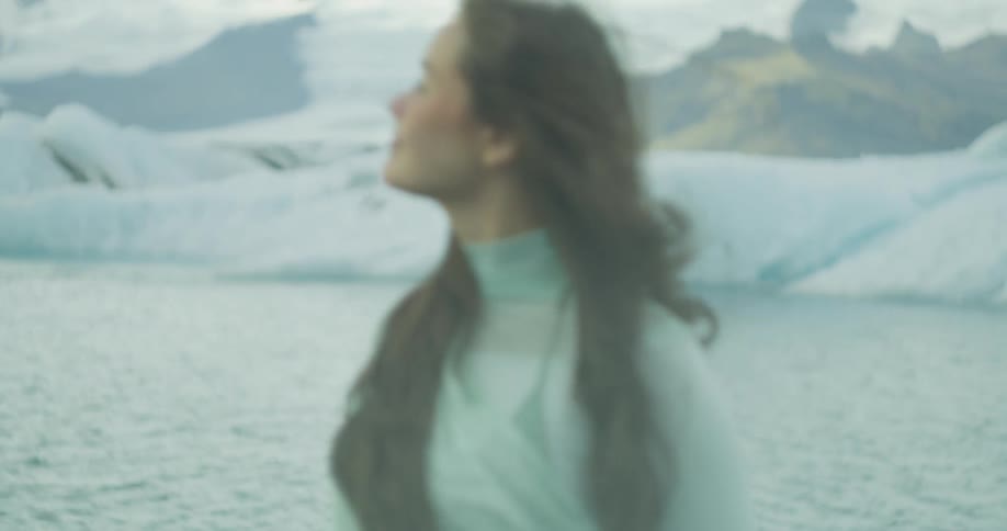 A pregnant woman in a flowing green dress gently holds her belly while standing before an icy glacial lagoon