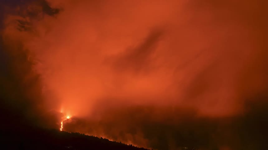 A terrifying nighttime view of a massive forest fire engulfing a mountainside, with thick smoke turning the sky a hellish orange