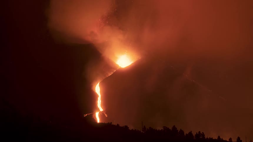A volcano erupts violently at night, spewing ash and smoke into the air as rivers of molten lava flow down its slopes