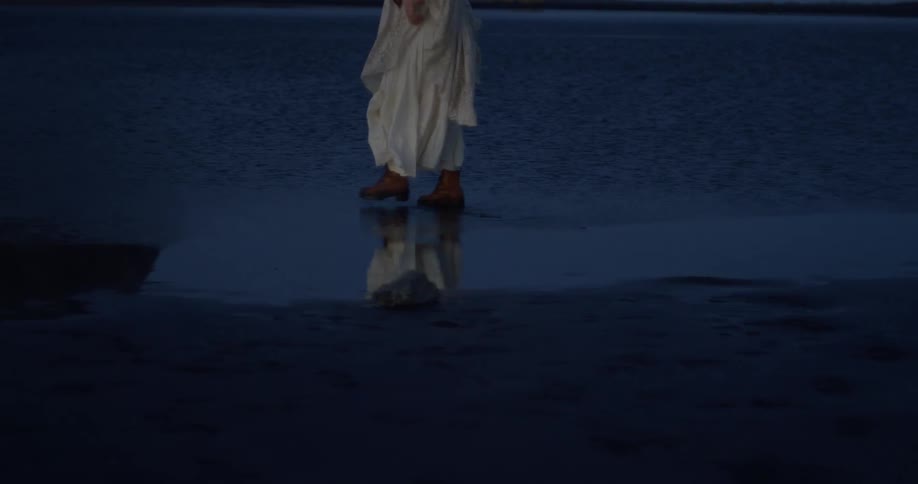 A mysterious figure in a flowing white dress and boots slowly walks through shallow, reflective water at dusk