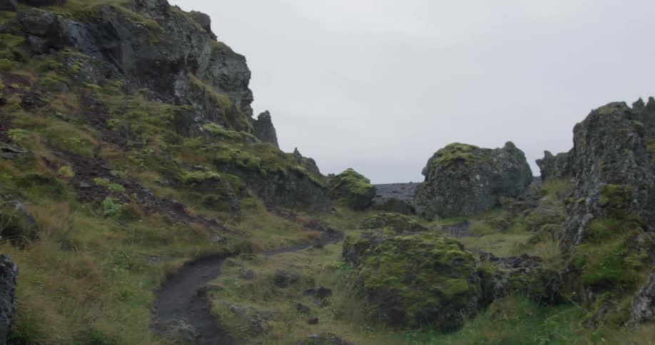A slow pan reveals a narrow dirt path winding through a rugged, moss-covered volcanic rock landscape on an overcast day