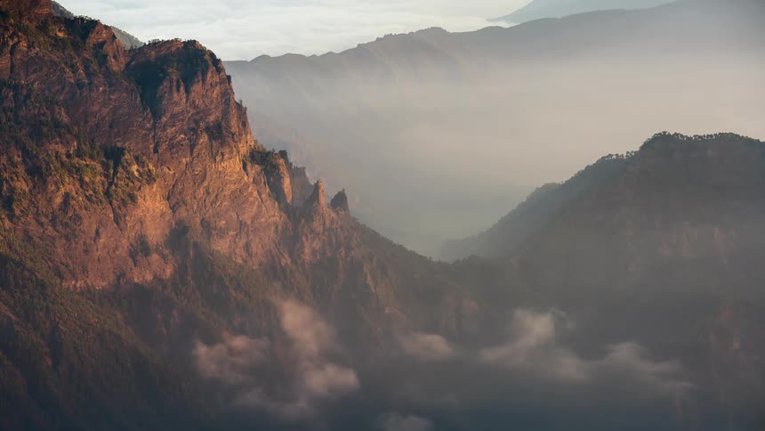 Golden sunrise light illuminates the rugged mountain peaks as clouds flow like a river through the valley below