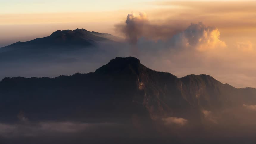 A volcano emits a plume of dark smoke over a serene mountain range shrouded in fog during a beautiful sunset
