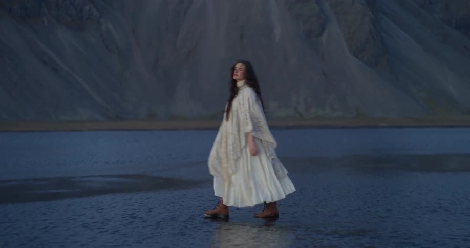 A woman in a flowing white dress and lace shawl walks pensively through the shallow water of a moody Icelandic landscape at twilight