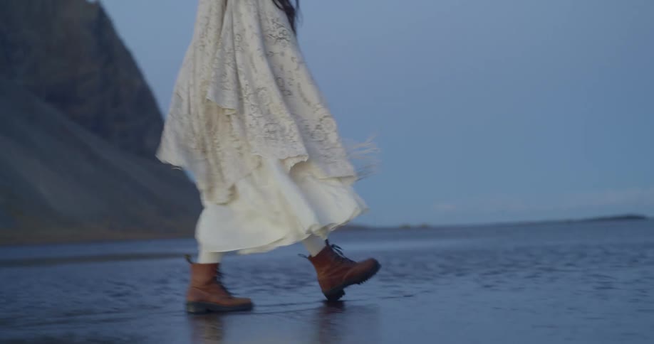 An ethereal woman in a flowing white dress and boots walks through the shallow waters of a remote beach in front of a majestic mountain at dusk