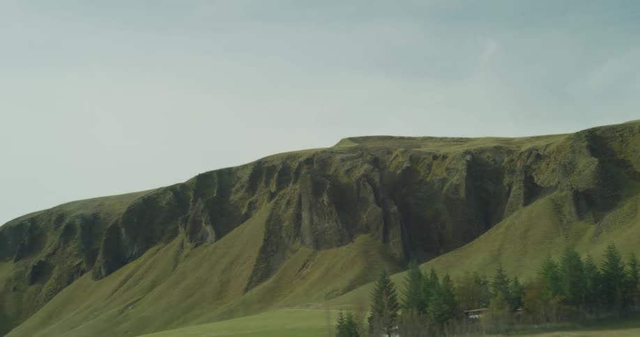 A scenic driving shot reveals the vast, mossy green hills and rugged cliffs of a dramatic Icelandic landscape