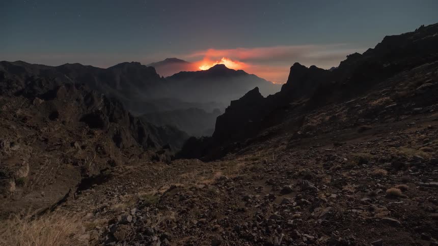 A stunning night time-lapse captures a fiery volcanic eruption in a vast, rocky mountain range under a starry sky