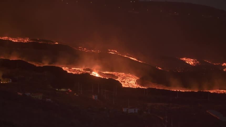 Rivers of molten lava from an active volcano flow down a dark hillside at night, threatening a small town below