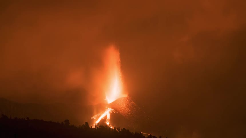 A volcano erupts at night, spewing fiery lava high into the sky and creating a dramatic display of smoke and ash