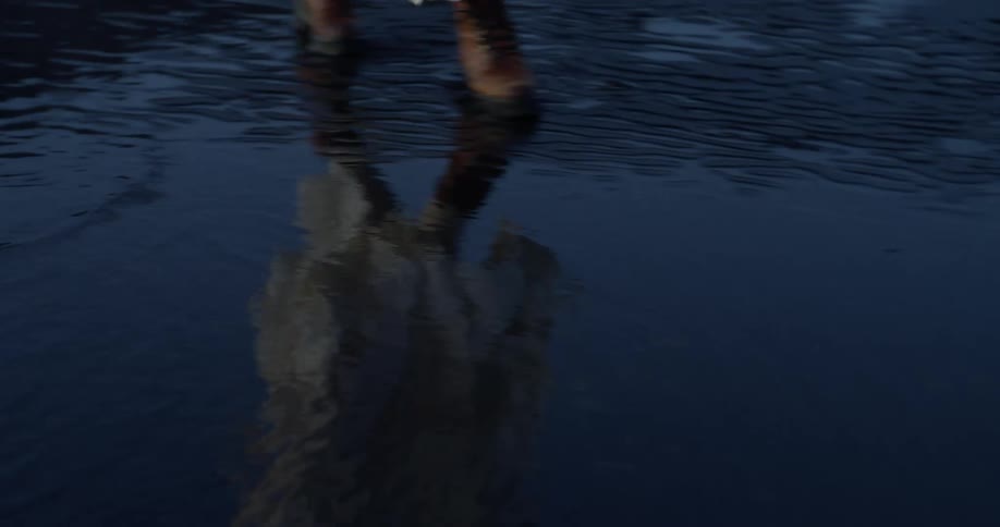 A close-up of a woman in a flowing white dress and heavy boots walking through the dark, shallow waters of a beach, her reflection rippling below