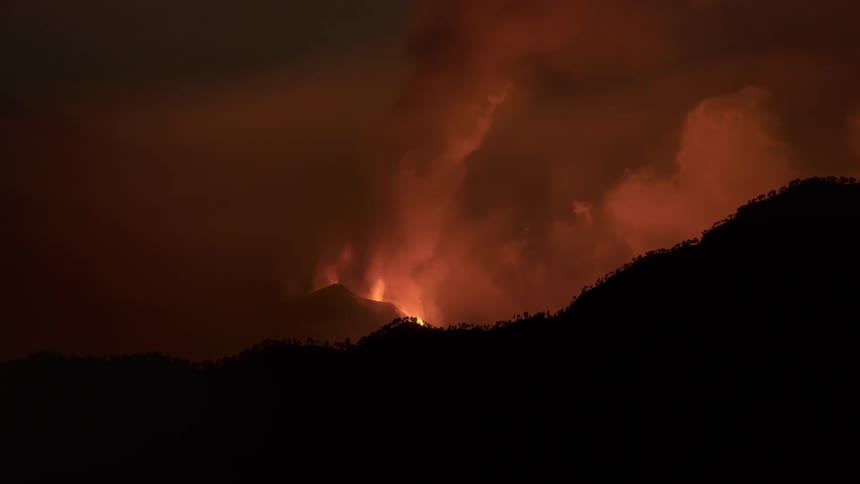 A raging wildfire burns on a distant mountainside at night, illuminating the smoky sky with a terrifying orange glow