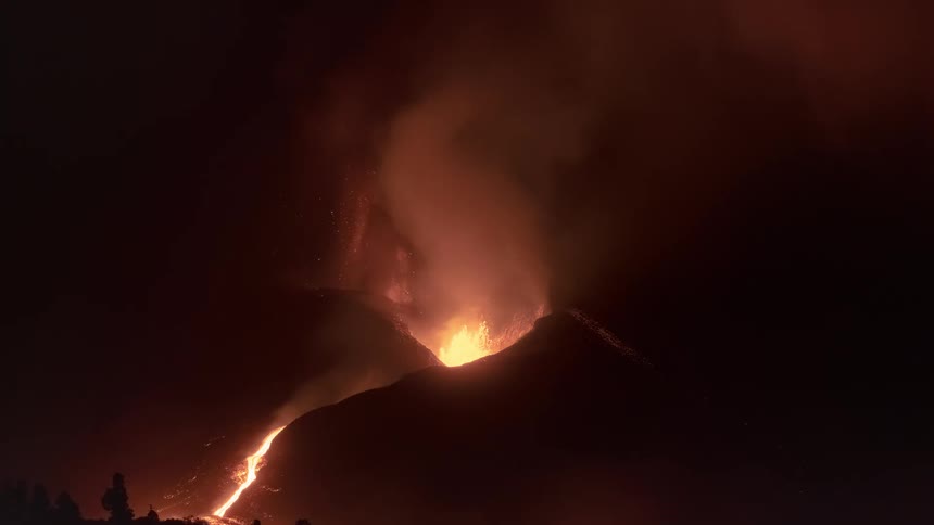 A volcano violently erupts at night, spewing glowing lava and thick smoke into the dark sky as molten rock flows down its side