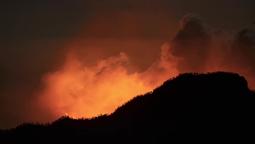 A silhouetted mountain range at dusk with a powerful volcanic eruption glowing fiercely in the background