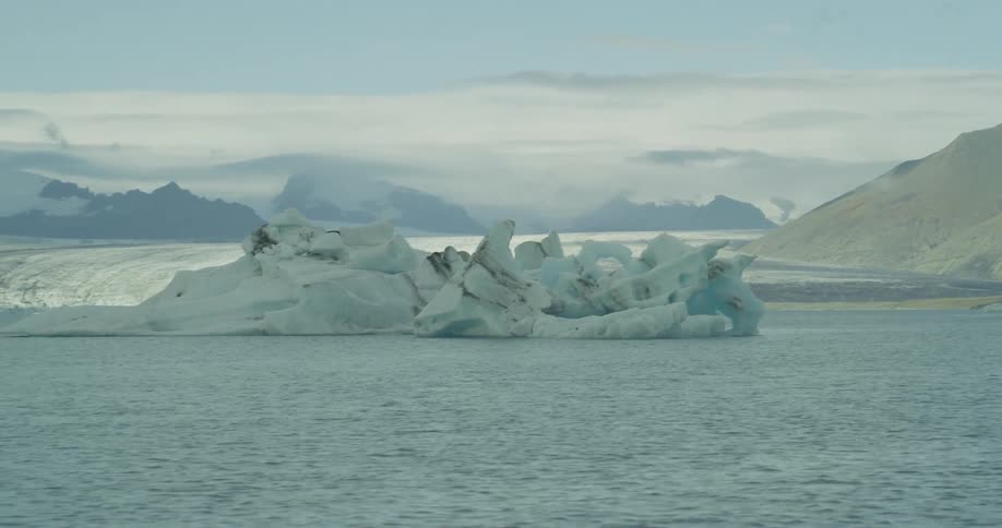 A slow pan reveals massive icebergs floating in a calm glacial lagoon with a sprawling glacier and mountains in the background