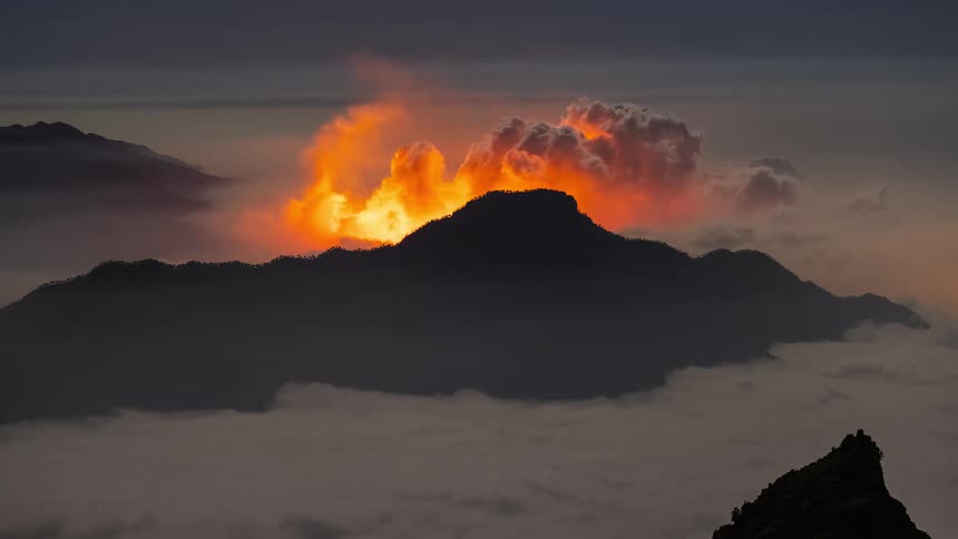 Dramatic fiery clouds resembling a volcanic eruption billow over a mountain peak at night above a sea of fog