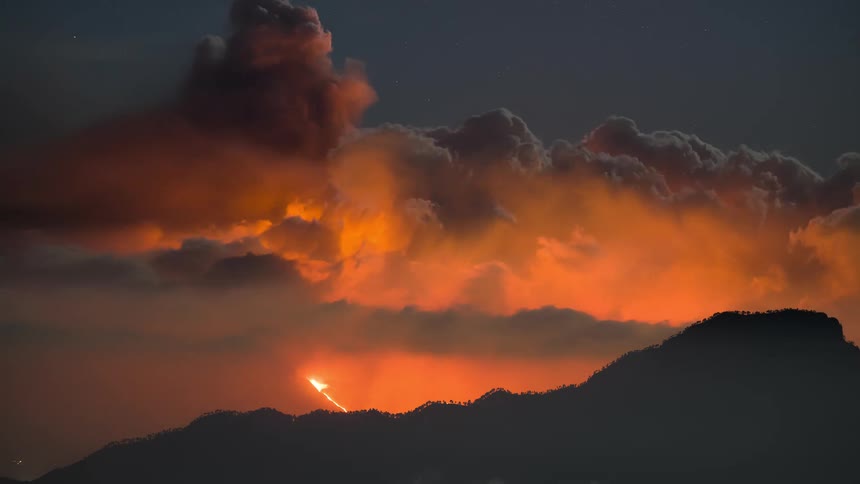 A dramatic time-lapse of a volcano erupting at night, with fiery orange clouds and lightning striking above a mountain silhouette