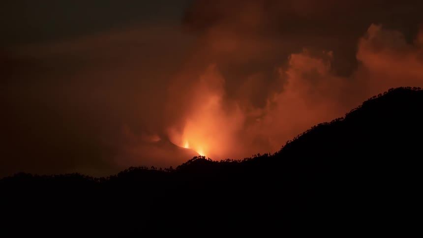 Dramatic night sky filled with fiery orange clouds and smoke from a volcano erupting behind a silhouetted mountain