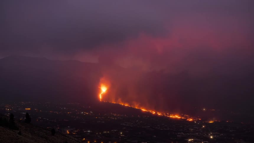 A powerful nighttime timelapse shows a volcano erupting, with glowing lava flowing down the mountain and a massive red smoke plume filling the sky above a town