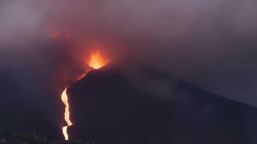 A dramatic time-lapse captures a volcano erupting through a thick cloud of smoke, with fiery lava flowing down its side and illuminating the sky