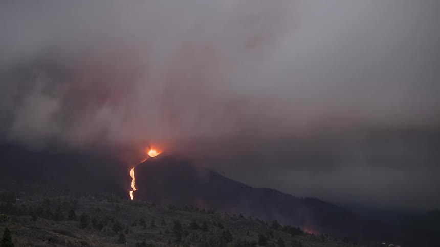 A dramatic time-lapse shows a volcano erupting at dusk, with rivers of lava flowing down its slopes and an intense fiery glow illuminating the billowing smoke and clouds
