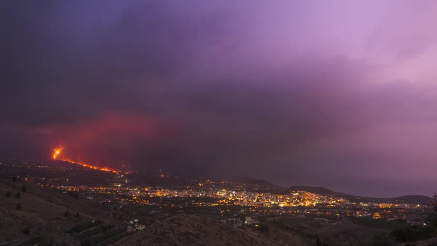 Time-lapse of the Cumbre Vieja volcano erupting on La Palma, with glowing ash clouds and lava flows threatening the city of Los Llanos below as day turns to night