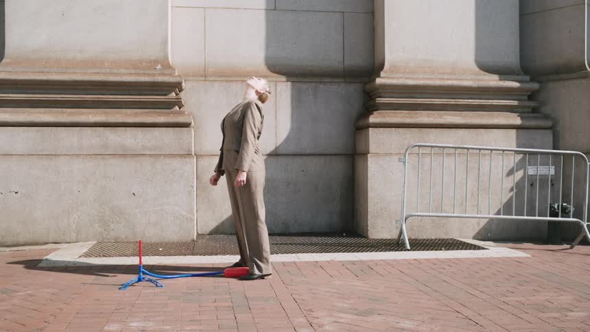 A businesswoman in a suit launches a toy rocket in slow motion on a city sidewalk before walking away