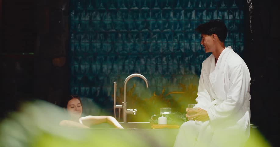 A romantic couple enjoys a relaxing evening at a luxury spa, with the woman in a steamy bathtub and the man in a bathrobe sitting beside her